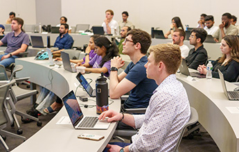 Students seated in a modern classroom with curved desks and open laptops.