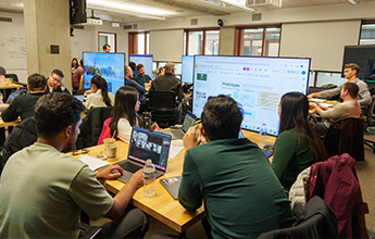 A collaborative workspace with people working on laptops around tables, featuring large display screens and a concrete column in the room.