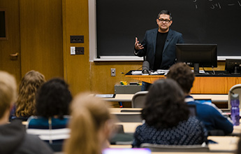 A lecturer speaking to students in a classroom with a chalkboard and computer
