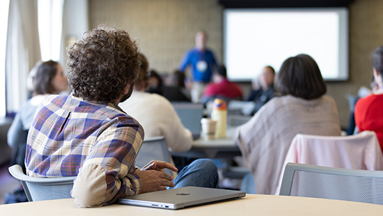 A classroom with people seated at tables facing a speaker in front of a large screen.
