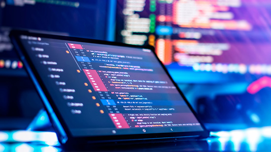 A tablet displaying a code editor with colorful syntax highlighting, set against a background of blurred code on a monitor.