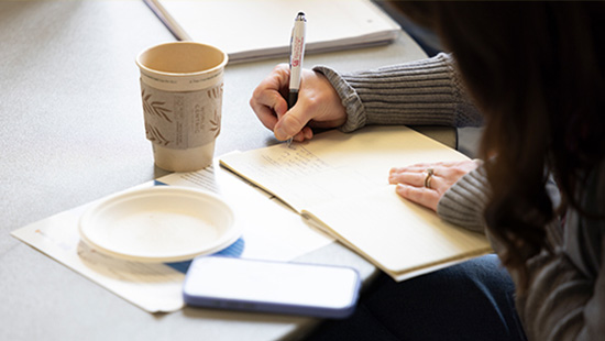 Person writing in a notebook with a cup of coffee and smartphone nearby.